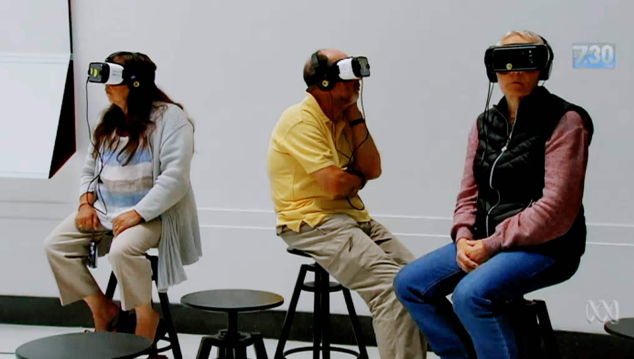 Three white people, a woman and two men, sitting on stools with virtual reality goggles covering their eyes. Each person is staring in a different direction. Video still.