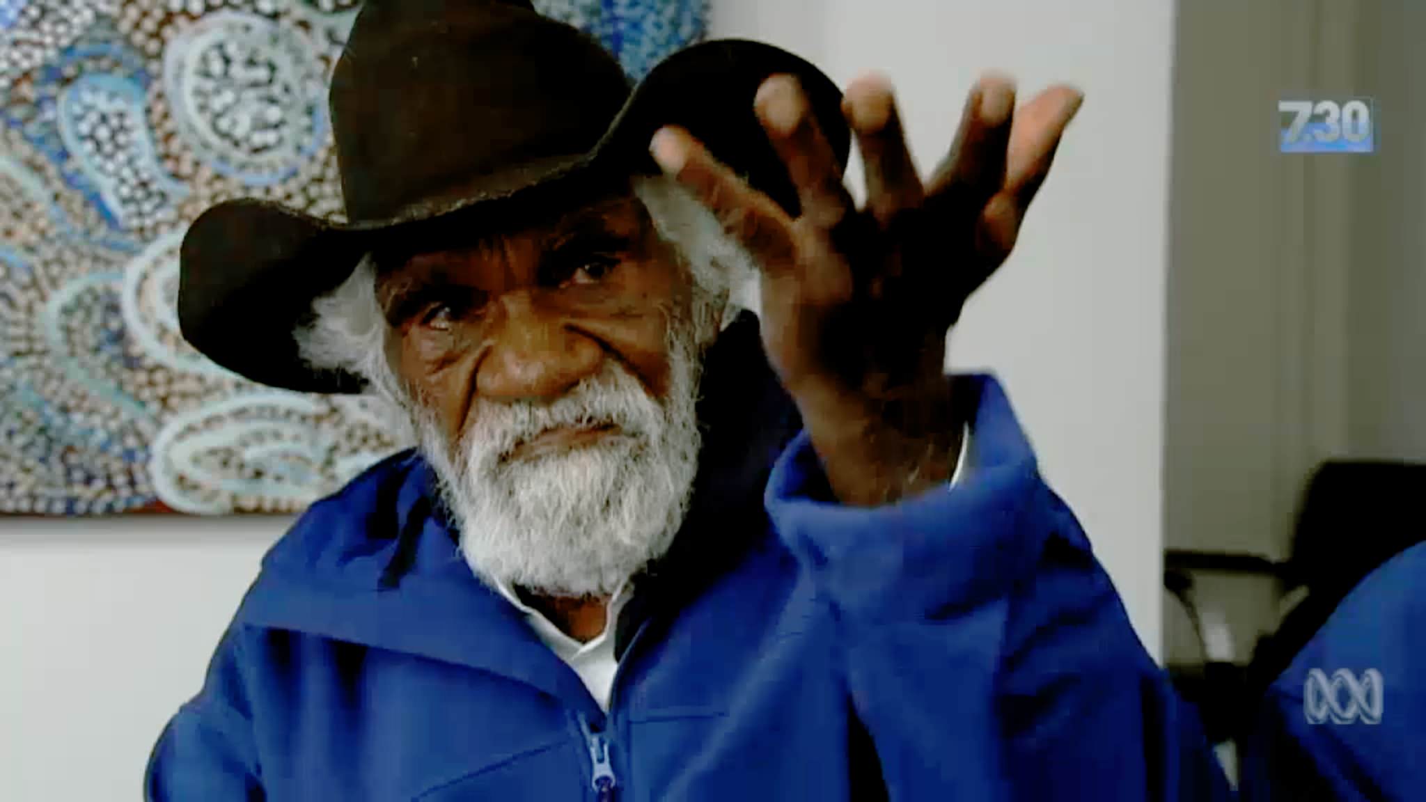 An older aboriginal man with wrinkled brown skin and white beard and hair wearing a cowboy hat and a light blue jacket. He is holding an open hand up to the sky and staring at the viewer with a serious expression. Video still.