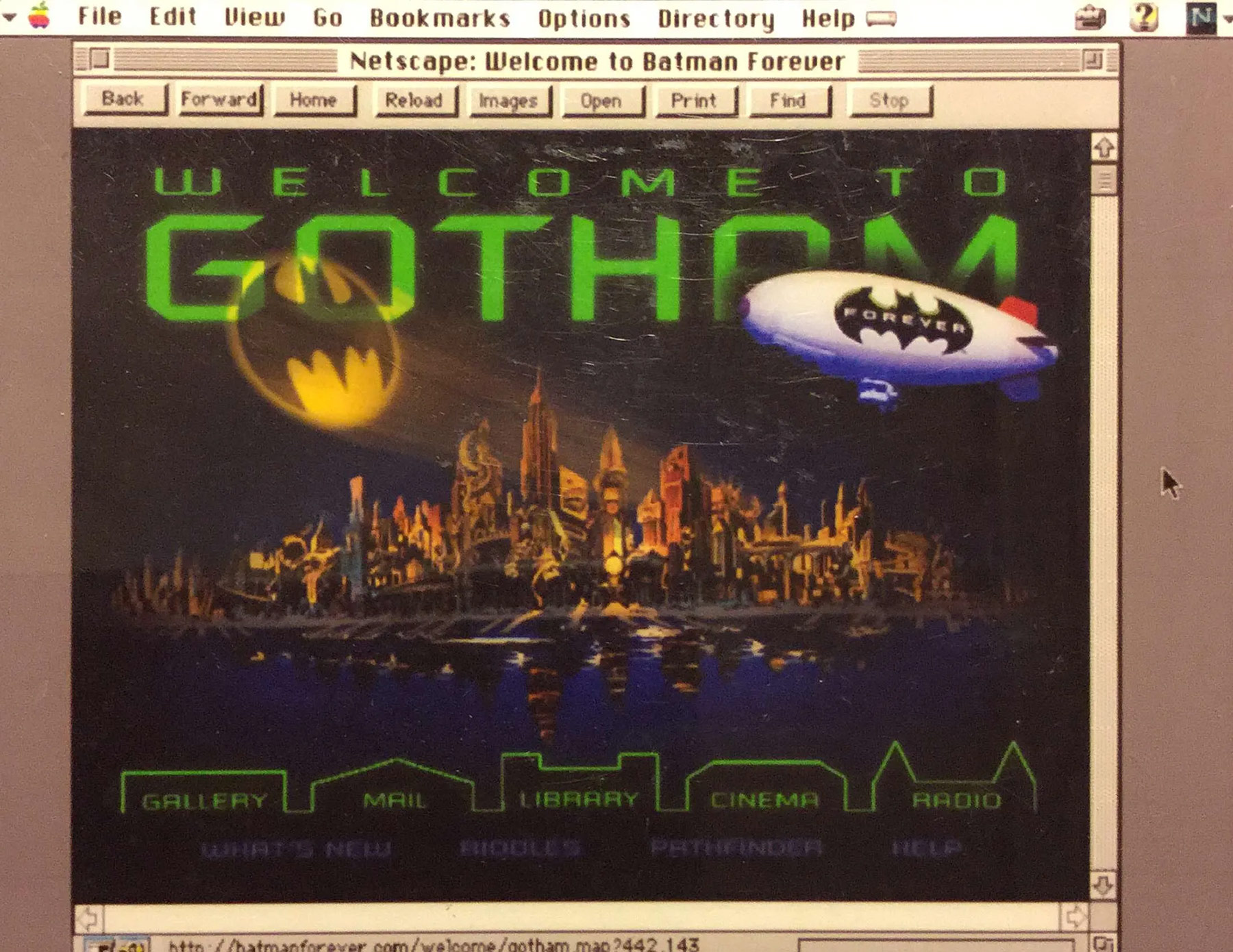 A slightly blurry photograph of the Batman Forever website, being displayed on the Netscape web browser. A headline set in a lime green scifi font reads, 'Welcome to Gotham'. Below the headline is a gothic noir cityscape at night, with the Batsignal reaching out into the night. A blimp is also present, with the Batman logo and the text, 'Forever' on its side. Below the skyline is more navigation options.