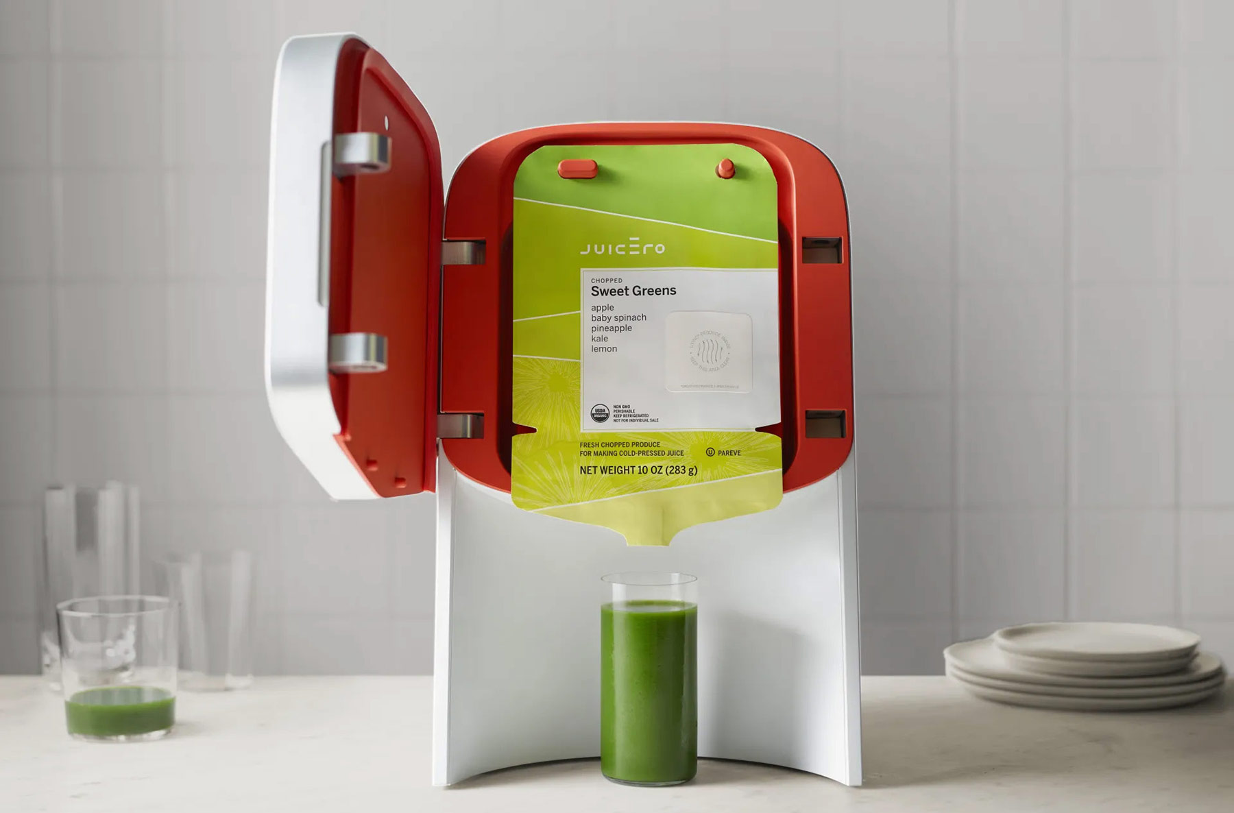The Juicero, a smoothly machined aluminum device sitting on a spotless kitchen counter. Its front plate is swung open, showing a branded juice packet with a chopped sweet greens flavor. Below the packet is a tall glass of green juice.