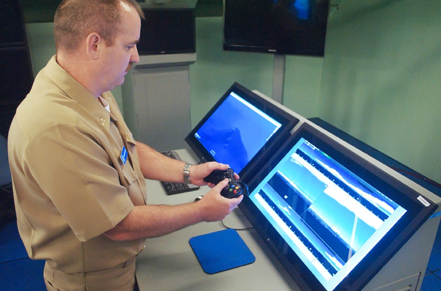 A man in uniform holding an Xbox controller. He is standing over two large displays, one showing a simulated seascape.
