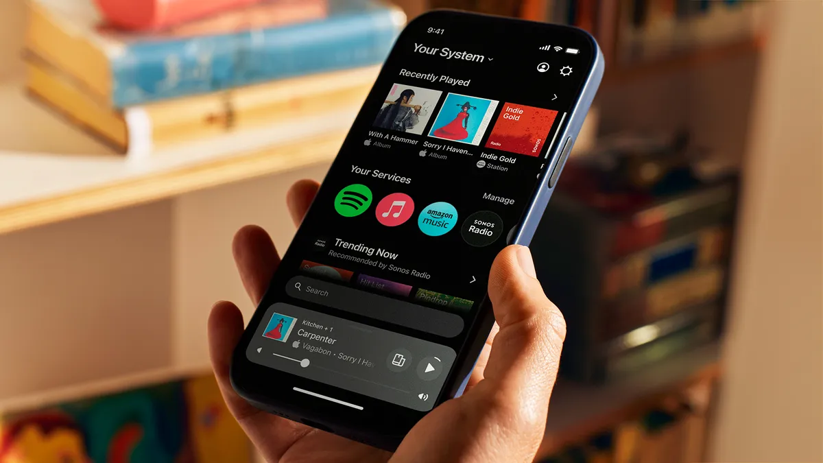 A promotional photo for Sonos' app. A hand is holding an iPhone that displays the Sonos app. The app is playing the song 'Carpenter' by Vagabon. It is also displaying Recently played songs, connected services, and trending music. The background is a warm-feeling living room, set to soft focus.