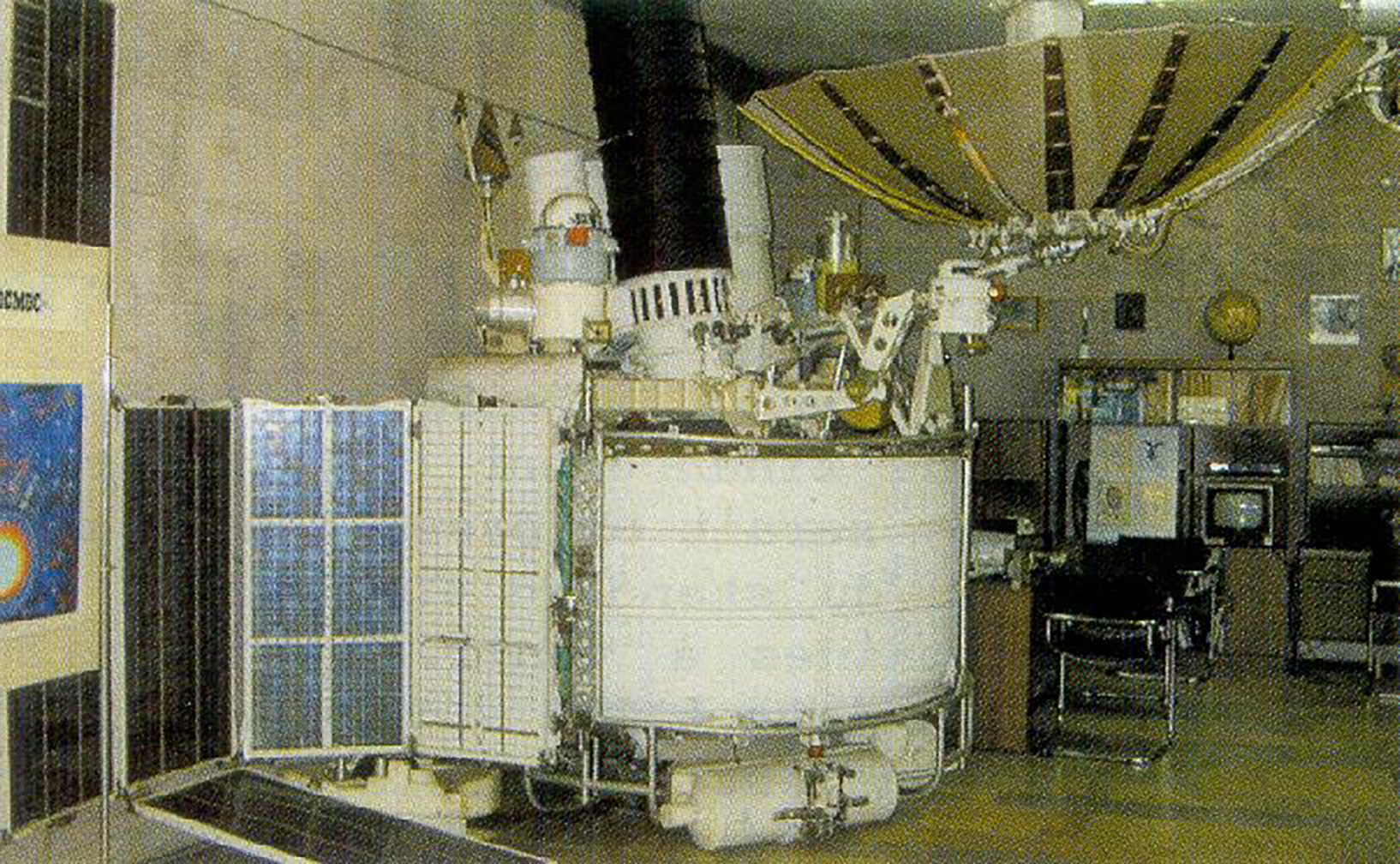 A grainy photograph of a white, drum-shaped satellite sitting inside a concrete office. It has a large golden, upwards-pointing dish in the shape of an umbrella, and a segmented solar panel wing. Carious structures and impliments also adorn the top of the drum.